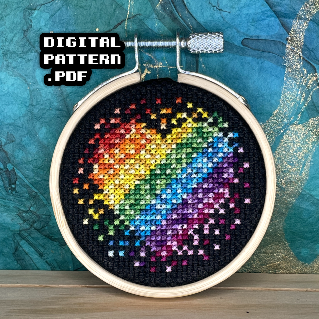 Picture of a completed cross stitch on black aida cloth in a tiny round wooden hoop of a heart in a diagonal rainbow gradient. The gradient starts with red on the top left, to purple on the bottom right. Digital Pattern PDF is in bold text in the top left.